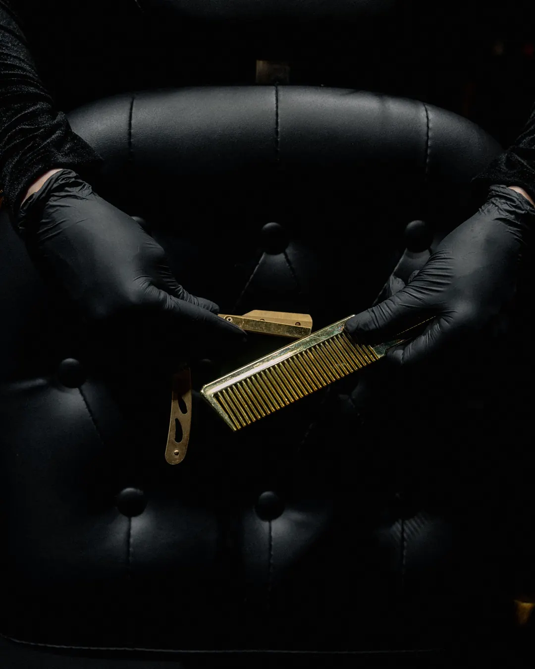Mains gantées de noir tenant un peigne doré devant un fauteuil capitonné noir